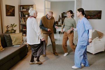 Group of two senior Caucasian women, senior Caucasian man and young adult Hispanic woman laughing and dancing together in living room, nurse wearing medical scrubs joining activity