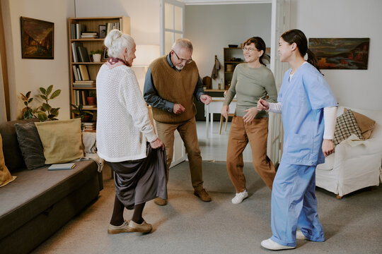 Group of two senior Caucasian women, senior Caucasian man and young adult Hispanic woman laughing and dancing together in living room, nurse wearing medical scrubs joining activity - Powered by Adobe
