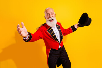 Bearded elderly circus magician in red costume extends arms welcomes audience with joyful smile...