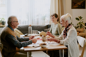 Senior Caucasian man and senior Caucasian woman sitting at table playing cards with middle aged Caucasian woman smiling in background, group enjoying leisure activity indoors