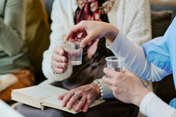 Senior Caucasian woman sitting with open book receiving plastic cup of water from young adult Caucasian woman in medical uniform, hands visible, focus on caregiving interaction