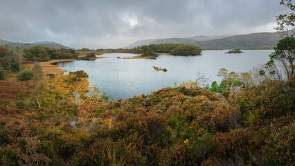 Panoramic view of a remote Kerry lake with autumn bogland, heather, islands, and misty mountains under a cloudy sky.
