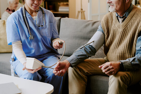 Caucasian senior man sitting on sofa having blood pressure measured by young adult Caucasian female nurse wearing stethoscope, nurse smiling while interacting with patient