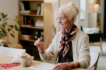 Senior Caucasian woman sitting at table concentrating on playing bingo game, holding bingo chip in hand, wearing jewelry and scarf, cup and napkin on table, bookshelf in background