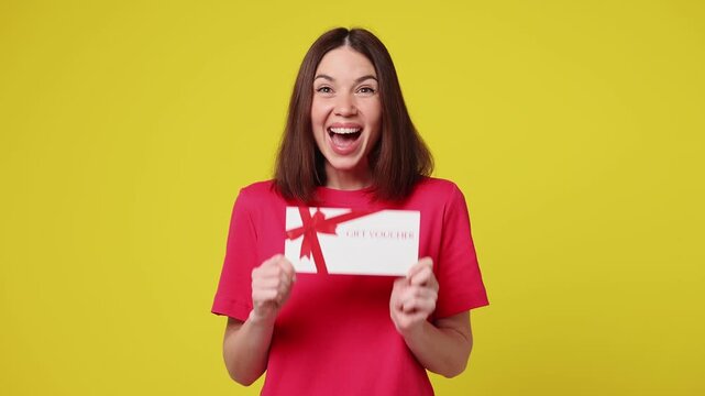 Surprised shocked happy young woman wear pink t-shirt casual clothes pointing finger on gift certificate coupon voucher card for store doing winner gesture isolated on plain yellow color background