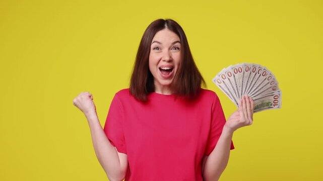 Happy young woman wear pink t-shirt casual clothes hold in hand fan of cash money in dollar banknotes doing winner gesture celebrate isolated on plain yellow color background studio. Lifestyle concept