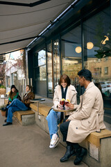 Couple sharing warm drinks outside modern cafe during cold autumn afternoon. Concept of seasonal beverage promotion, cozy date storytelling, hospitality branding and atmosphere visuals