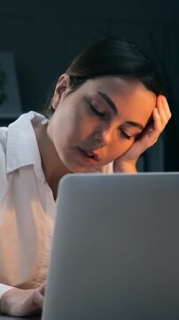 Exhausted young businesswoman leaning on hand and struggling to stay awake while working on laptop computer late at night. Overworked female employee. Vertical video.