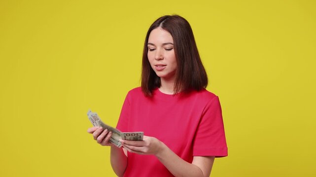 Happy accountant young woman wear pink t-shirt casual clothes hold in hand count stack of cash money in dollar banknotes isolated on plain yellow color background studio portrait. Lifestyle concept