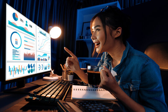 Smiling young beautiful asian creative looking on pc of profit of sales target report and writing on notebook for memo with planning invest new strategy market project at neon light night. Stratagem.