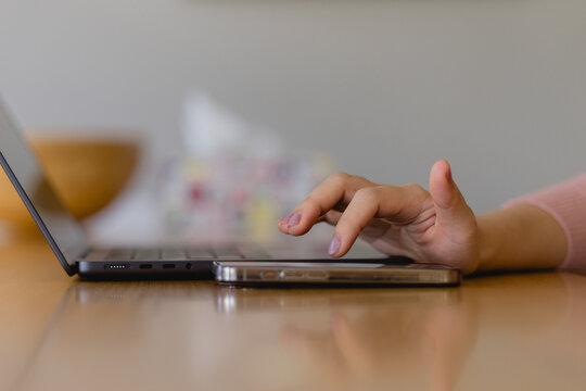 woman’s hand touching a smartphone beside a laptop on a wooden desk, representing multitasking, digital communication, remote work modern tech lifestyle.
