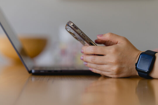 woman’s hand touching a smartphone beside a laptop on a wooden desk, representing multitasking, digital communication, remote work modern tech lifestyle.