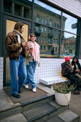 Friends chatting with warm drinks near modern cafe on autumn day. Concept of urban lifestyle ads, seasonal communication, event spot ambience, and gentle wellbeing storytelling.