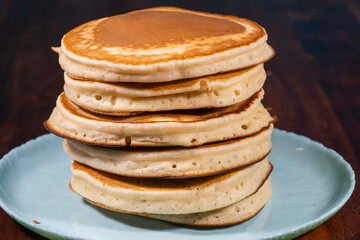 A high stack of six golden-brown, fluffy pancakes is centered on a light blue plate against a dark background