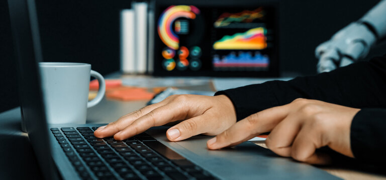 A focused work environment featuring hands typing on a laptop, accompanied by business graphs on a monitor, a coffee cup, and a robotic arm in the background.Noogenesis