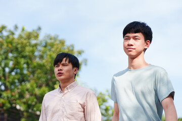Two Asian men, father and teenage son, walking together side by side in the park in afternoon on sunny day with green tree, blue sky in background. Exercise routine, family healthy lifestyle concept