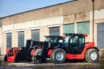 Two black and red telehandlers versatile agricultural and construction vehicles with attachments such as a lifting basket and a large bucket or scoop parked in concrete machinery yard.