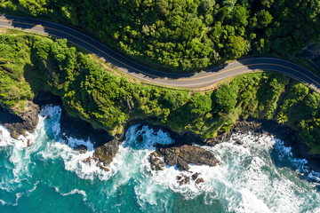 Aerial top view of winding coastal road with green forest and ocean waves crashing
