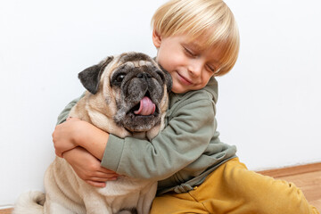 A little boy hugs a funny dog pug with closed eyes and yawning. The image symbolizes love, trust, friendship and sincere emotional connection between child and pet.
