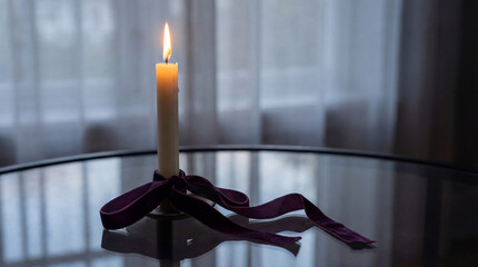 Single burning candle with purple ribbon on reflective glass table in dim room with sheer curtains and soft natural light