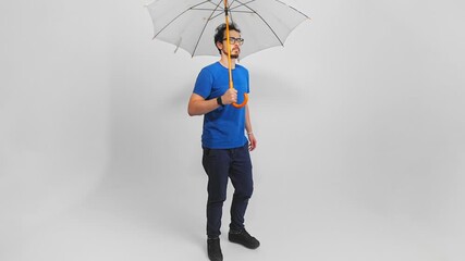 Confident young adult Caucasian man with a beard and glasses walks slowly holding umbrella above him. Relaxed guy in casual clothes is moving in changing atmospheric conditions filmed in studio