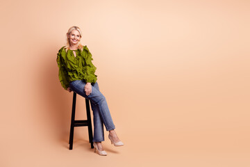 Confident mature woman with blonde hair wearing a green ruffled blouse and jeans sits on a stool in...