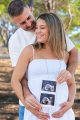Pregnant couple holding ultrasound photo and smiling outdoors with love. Expecting parents showing...