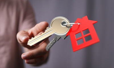 Close-up shot of a person's hand offering house keys with a red house-shaped keychain. Concept of homeownership, real estate, buying a house.
