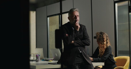 Senior businessman standing with thoughtful pose in office boardroom while coworkers concentrate on laptops during company meeting