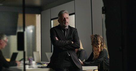 Senior businessman standing with thoughtful pose in office boardroom while coworkers concentrate on laptops during company meeting