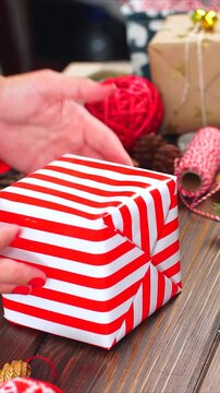 vertical video of female designer hands packing Christmas presents in wrapping papers on the wooden table with decorating items and Christmas baubles