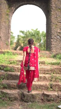 A young woman wearing a traditional red salwar kameez with golden embroidery stands outdoors near an old stone wall covered with natural moss.