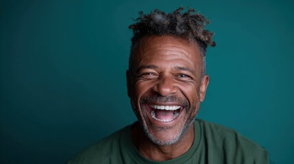 A full of life and cheerful man with wavy hair smiling brightly against a teal background, radiating warmth, happiness, and positivity in a relaxed setting.