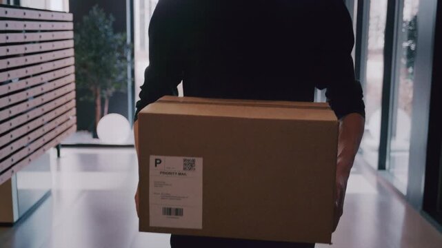 Anonymous courier walking through a modern building lobby carrying a cardboard package for delivery