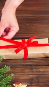 Hands wrapping brown gift with ribbon, close-up of hands with red nails tying neat red bow on kraft paper-wrapped parcel on rustic wooden table, scissors and pine sprig accents, vertical video