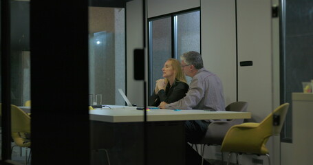 Senior coworkers talk face to face across office desk with laptop and documents, analyzing work updates together in quiet corporate meeting environment