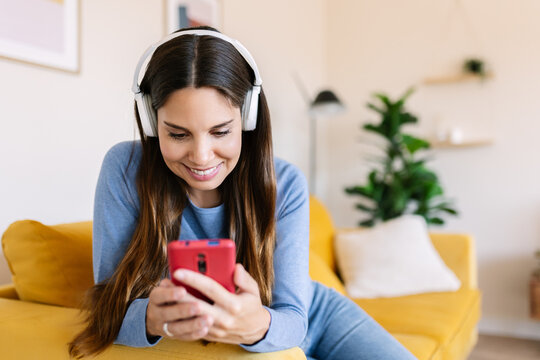 Smiling Caucasian woman using smartphone with headphones on sofa. Happy female enjoying music and connection at home. Technology and lifestyle concept.
