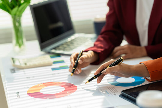Two businesswomen are holding and pointing at a financial report, discussing marketing strategies, pointing at the company's business report computer screen.