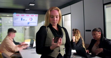 Serious businesswoman looks at smartphone with concern during office meeting, focused team working on laptops, data visualization screen in background