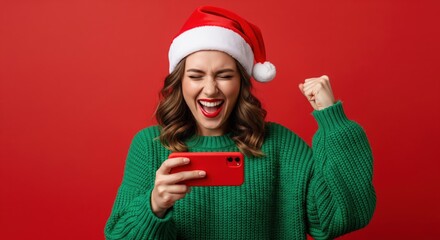 Excited young woman in Santa hat cheers while celebrating a victory on her smartphone against a red background.