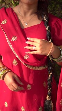 A young woman wearing a traditional red salwar kameez with golden  embroidery stands outdoors near an old stone wall covered with natural moss.