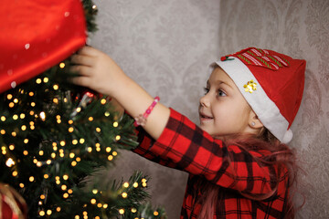 Smiling girl in plaid shirt and Santa hat hanging ornaments on a Christmas tree