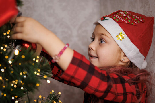 Little girl in a Santa hat decorating a Christmas tree, smiling and focused on ornaments.