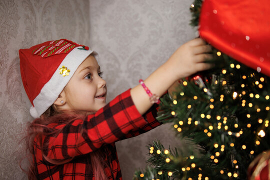 Little girl in a Santa hat decorating a Christmas tree, smiling and focused on ornaments.