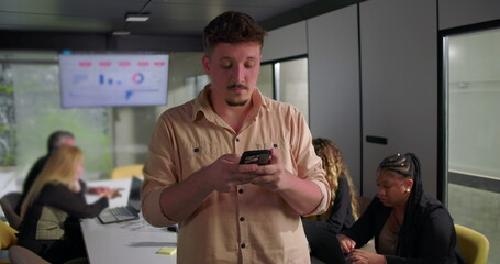 Serious young businessman checking smartphone during office meeting, colleagues working at laptops in modern conference room with analytics screen in background