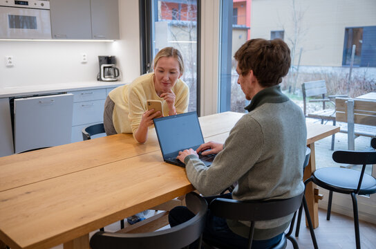 Woman showing smartphone to man working on laptop in modern kitchen