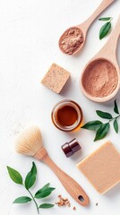 Overhead shot of various cosmetic products like soap, powder, brush, and oil on a white textured surface, surrounded by green leaves.