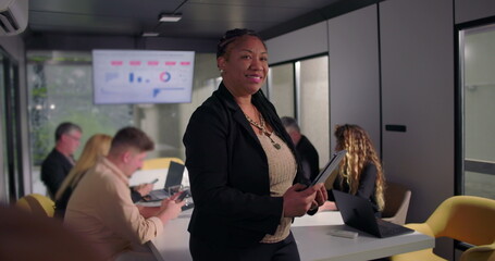 Smiling African American businesswoman holding digital tablet in office, confident leadership presence as team works at conference table in background