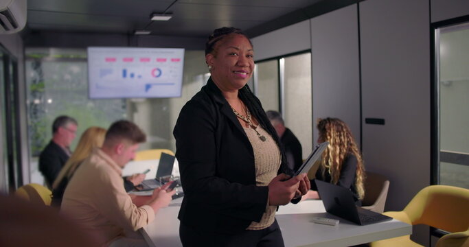 Smiling African American businesswoman holding digital tablet in office, confident leadership presence as team works at conference table in background - Powered by Adobe