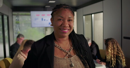Smiling African American businesswoman looking at camera in modern office, team in background working during meeting with analytics chart on digital display